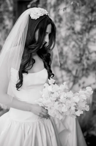 Bridal portrait of a bride holding a bouquet, eyes downcast, framed by garden foliage and a wall for a quiet, timeless feel.