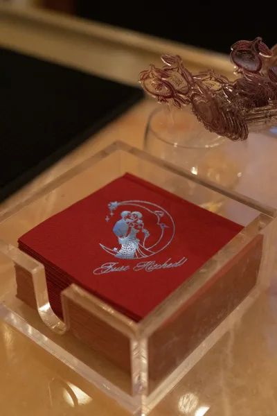 Custom wedding napkins, foil-stamped and stacked in an acrylic napkin holder on a bar counter beside a glass and bar mat.