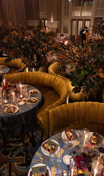 Reception tablescape with banquette reception seating and taper candles aglow in an indoor ballroom framed by tall windows and draped curtains.
