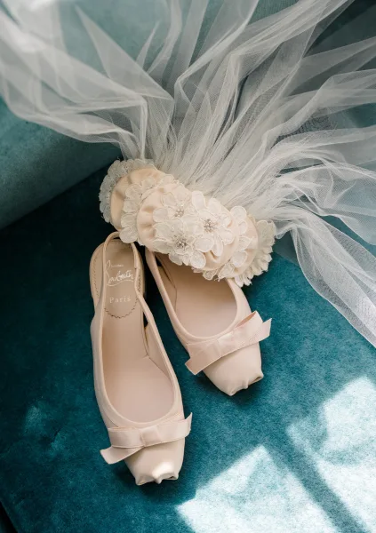 Bridal shoes, chic bridal flats with neat bows, rest on a teal velvet chair's plush seat, the rich hue framing their elegance.