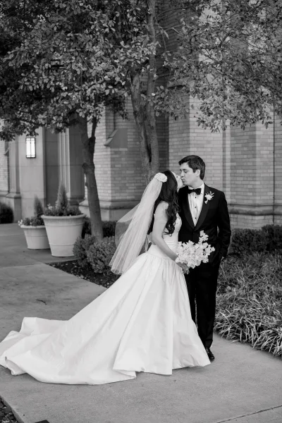 Wedding kiss portrait of a bride with long veil sharing a side-profile kiss with her groom under trees beside a brick wall.