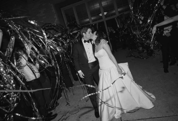 Wedding exit at a wedding streamer send off, the couple moves through guests waving streamer wands at nighttime beside a brick wall and large windows.