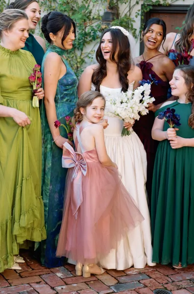Bridal party portrait with bridesmaids in mismatched dresses, the bride centered, bouquets in hand on a brick patio by a vine-covered wooden door.