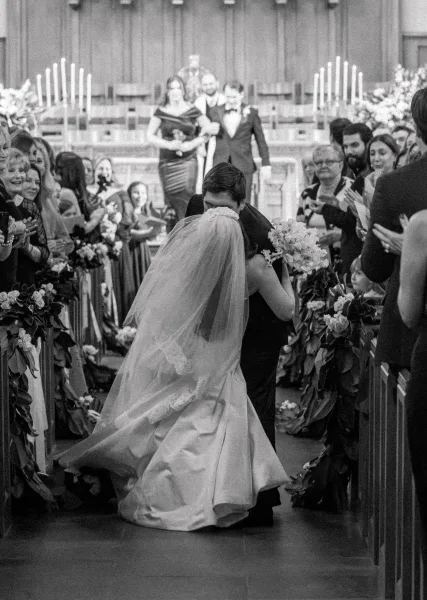 Recessional kiss during a ceremony exit moment, the bride clutching a bridal bouquet as they move down the church aisle, candlelit altar behind.