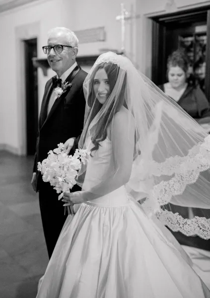 Father of the bride escorts his daughter at church, her lace veil catching light as they step through a doorway beneath a cross.