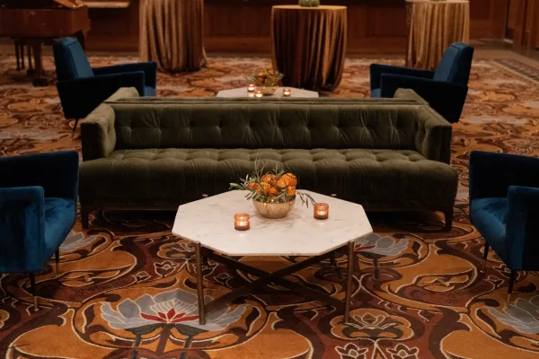 Wedding lounge seating styled as a reception lounge area, anchored by a velvet sofa, with a grand piano framed by wood paneling behind