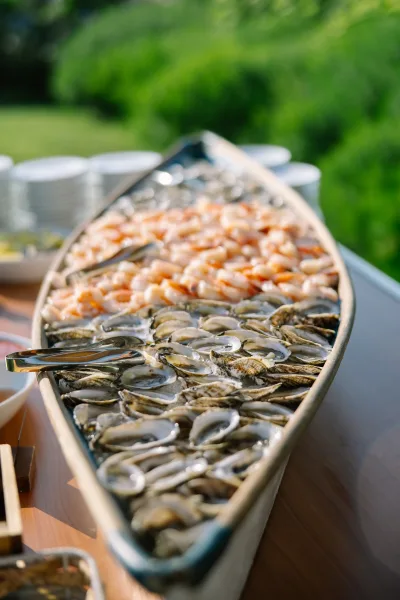 Raw bar display with oysters on ice arranged in a boat serving tray, set on a wood table amid garden greenery for an outdoor reception.