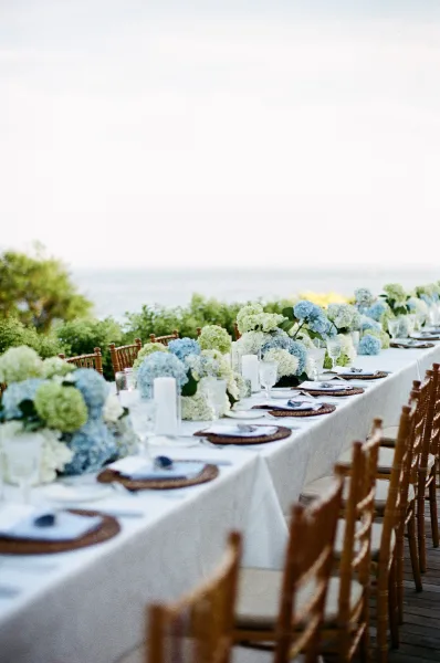 Reception tablescape for an outdoor reception table with hydrangea florals, set on a wooden deck overlooking an ocean view and garden greenery.