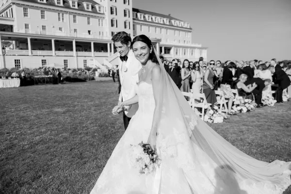 Ceremony recessional with bride and groom walking hand in hand, her cathedral veil flowing as they exit down the aisle on a hotel lawn.