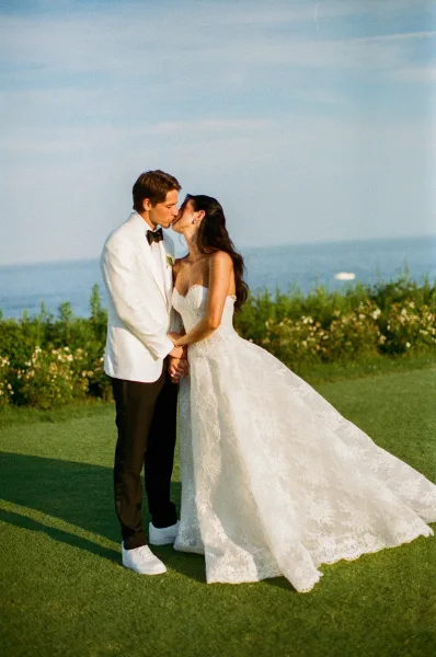 Wedding kiss portrait of a bride and groom holding hands by the ocean, her strapless lace gown glowing in golden hour light.
