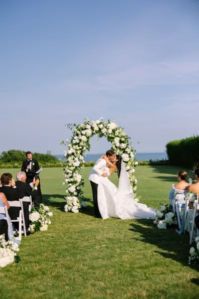 Ceremony kiss moment as the couple share their first kiss under a white floral arch on a grass lawn with an expansive ocean view.