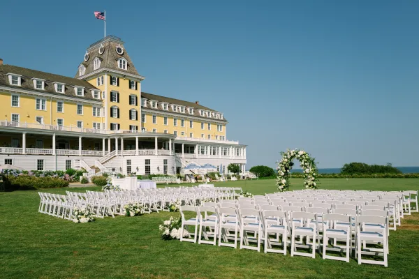 Outdoor ceremony setup with garden ceremony seating and a floral arch on a grand hotel lawn overlooking the ocean under a clear blue sky.