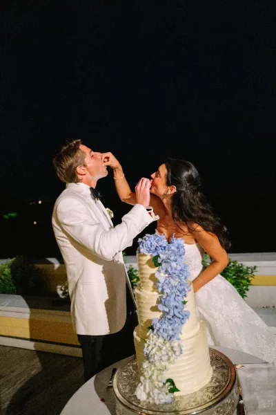 Cake cutting as the bride and groom feed each other cake beside a three-tier buttercream with hydrangea florals on an outdoor terrace at night.
