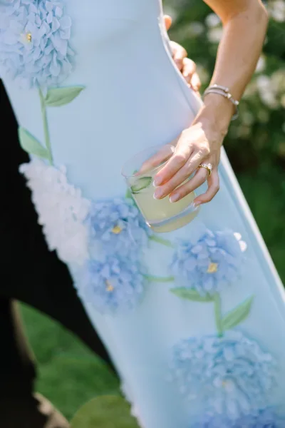 Dress detail of a blue floral dress, close-up showing a hand with a ring near the skirt, set against garden greenery and a grass lawn.
