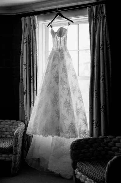 Wedding dress hanging by a bright window with delicate lace details, soft curtains behind and wicker chairs framing the scene.