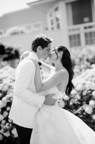 Wedding kiss portrait of a bride and groom, her bracelet catching light, as they embrace beside hydrangea bushes and a garden-side building.