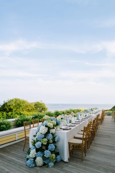 Reception tablescape on a long banquet table, with hydrangeas, set on a wood deck overlooking the ocean under a clear blue sky.