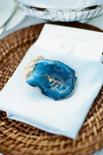 Oyster shell place card with gold calligraphy, styled as a coastal escort card, rests on a woven charger beside a simple paper card.