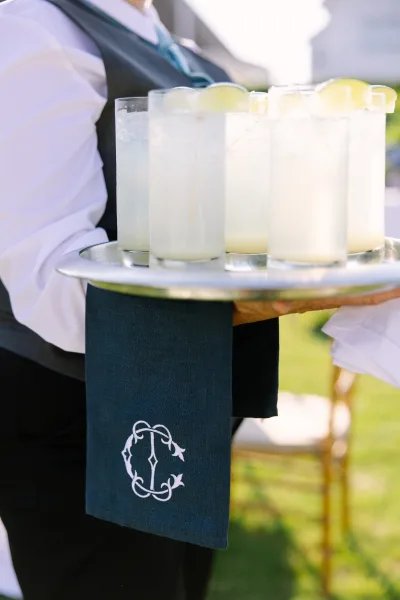 Wedding cocktails served via passed cocktail service on a silver tray, set against a sunlit grass lawn with a softly blurred chair in the background.