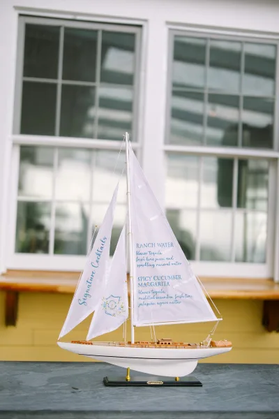 Signature cocktail sign for a wedding bar menu displayed on a model sailboat, set against window panes and a yellow wall on a tabletop.