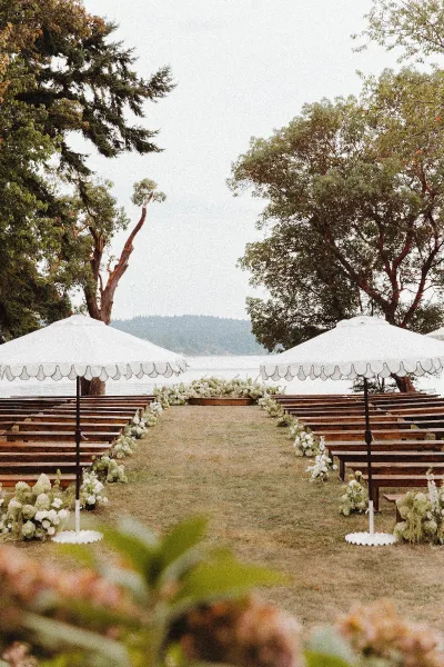 Ceremony setup with outdoor ceremony seating, wedding aisle flowers lining a grassy lawn with wooden benches and white umbrellas by the lake