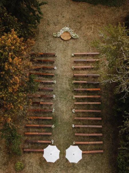 Ceremony setup for an outdoor wedding ceremony with wood benches flanking a round rug altar, floral accents and white umbrellas on a tree-lined lawn