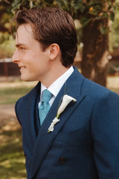 Groom portrait in a navy suit with patterned blue tie and white calla lily boutonniere, looking away in an outdoor garden setting