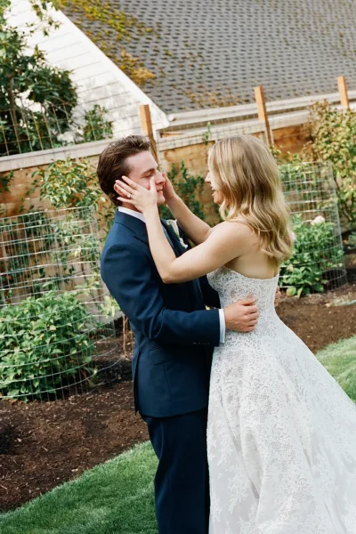 Couple portrait of bride and groom embrace, bride touching his face in a strapless lace gown beside garden greenery and fences