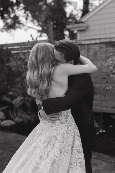 Wedding kiss as bride in a strapless lace dress and groom in a suit embrace on a garden lawn with trees and a wooden fence