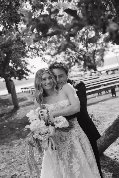 Couple portrait of bride and groom hugging, bride holding a bouquet in a strapless lace dress under trees by wooden benches outdoors
