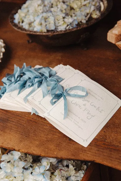 Wedding invitations arranged as a wedding invitation suite with calligraphy, tied with a blue ribbon on a wood table beside white flowers