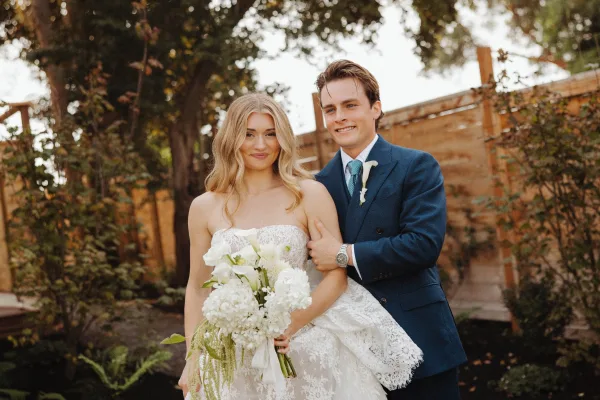Couple portrait of bride holding a white calla lily and hydrangea bouquet as groom hugs her in a garden by a wooden fence
