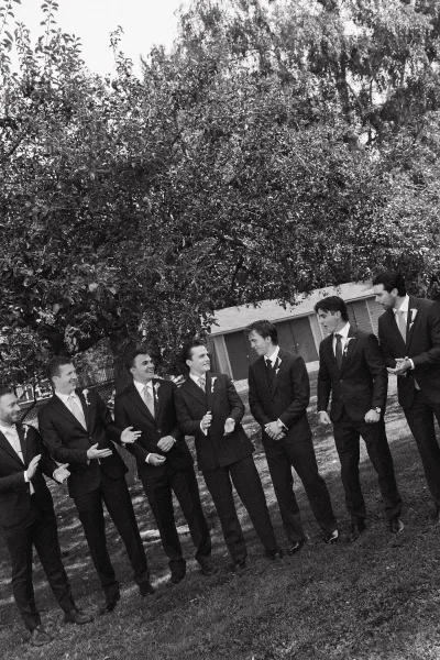 Groomsmen portrait of men in matching dark suits with boutonnieres standing in a row on a grassy lawn with trees behind