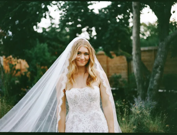 Bridal portrait of a smiling bride outdoors in a strapless lace wedding dress with a long veil, standing by garden greenery and a wooden fence