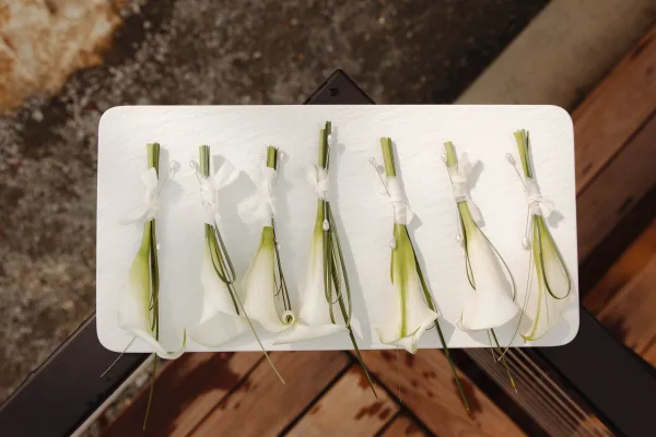 Boutonniere set of white calla lily boutonniere pieces with ribbon and greenery stems laid out on a white tabletop outdoors