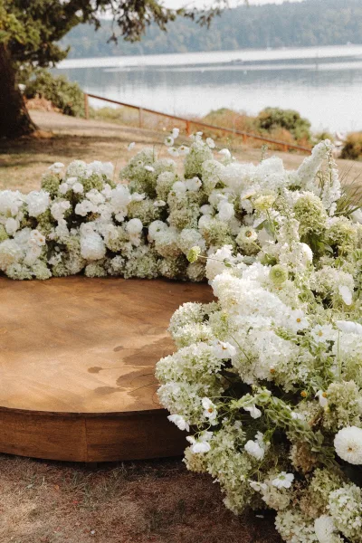 Ceremony floral arch with white hydrangeas on a round wooden platform, set on a grassy lawn by a lake with trees and hillside