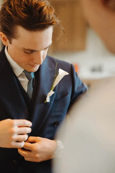 Groom portrait in a navy suit with a patterned tie, white calla lily boutonniere, and wristwatch against a softly blurred indoor reception backdrop