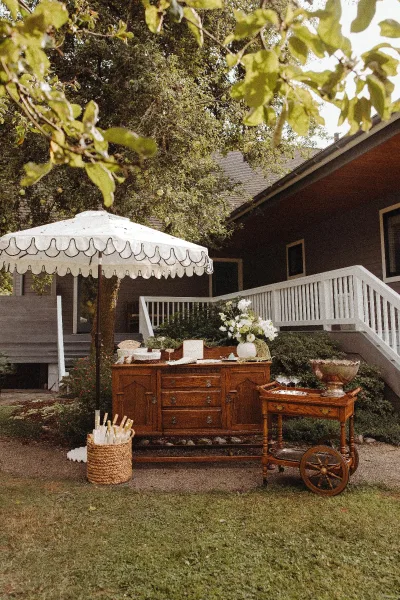 Wedding bar setup with a vintage wooden dresser and bar cart, glassware and ice bucket under a white patio umbrella on a porch