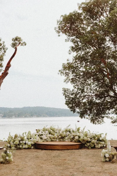Ceremony backdrop with a floral ceremony backdrop of white blooms forming a grounded arch on a round wooden platform by the lakeshore