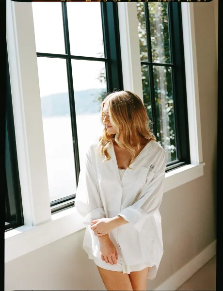 Bridal getting ready portrait of bride getting ready outfit in a white button-down shirt by black-framed windows with water view beyond