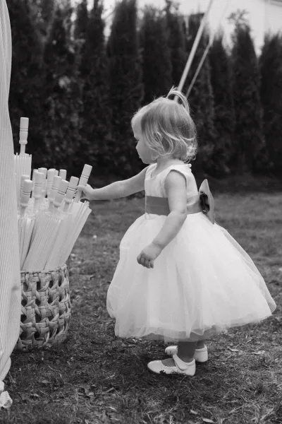 Flower girl moment as a toddler flower girl in a sash dress and Mary Jane shoes, holding a basket and parasols on a grassy lawn with trees