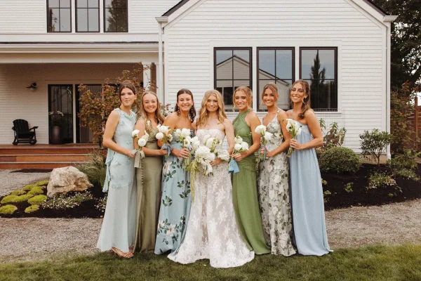 Bridesmaid group photo with bride with bridesmaids holding white bouquets with ribbon outside a white house porch and shrubs