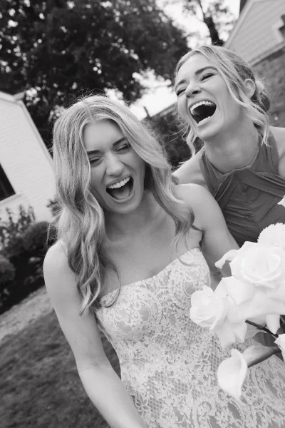 Bridesmaid portrait of a woman laughing while holding a white rose bouquet beside the bride on a lawn outside a house with trees