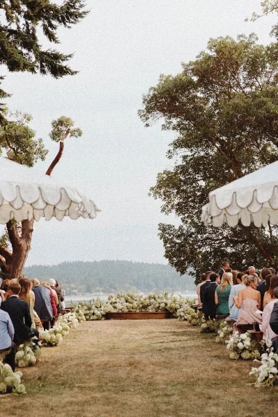 Ceremony aisle decor with white floral arrangements lining a grass lawn aisle, wooden benches and white parasols beside a waterfront view