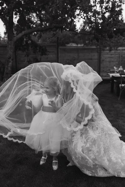 Bride with flower girl sharing a flower girl with veil moment, long lace wedding dress and veil on an outdoor lawn by trees and fence