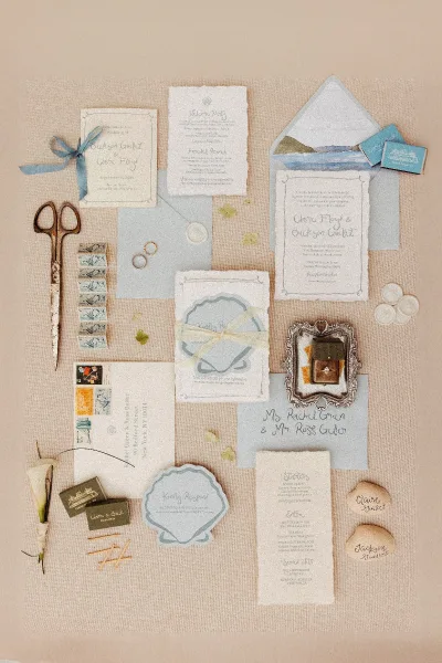 Invitation suite wedding stationery flat lay with dusty blue ribbon bow, wax seals, stamps, and rings on a linen fabric backdrop