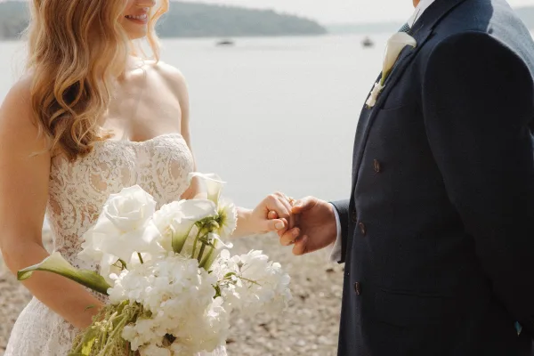 Couple portrait of bride and groom holding hands, showing wedding rings, on a beach shoreline with ocean and distant hills behind them