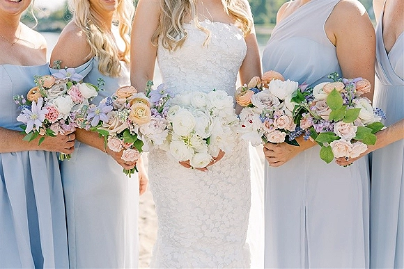 Bridesmaid bouquets in pastel bridesmaid bouquets palette with blush roses, white peonies and lavender, held by dresses near water outdoors