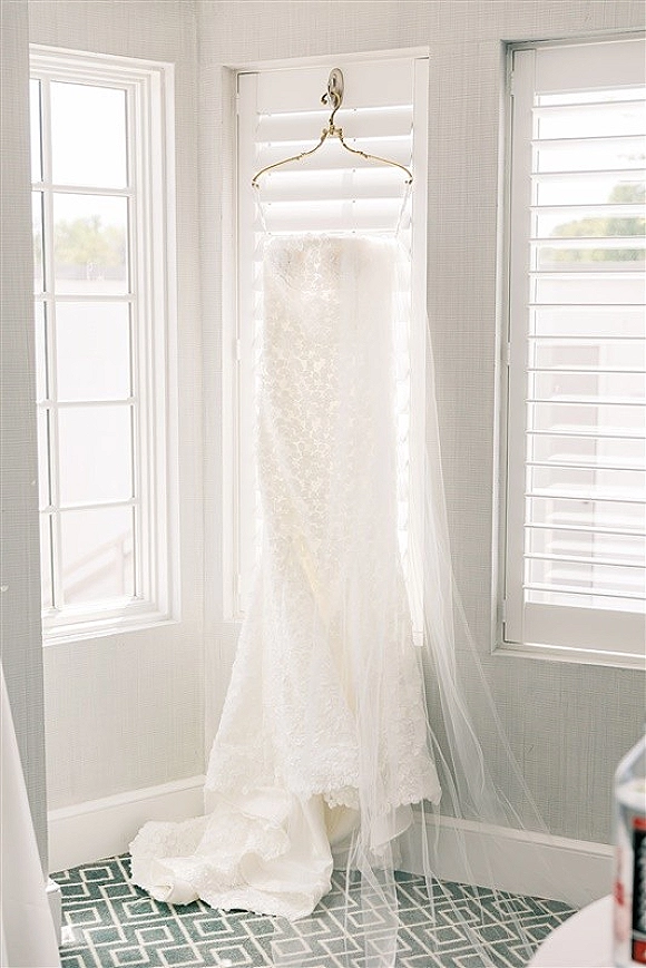 Wedding dress hanging by bright windows, lace bridal gown with tulle veil on a hanger against white shutters and patterned tile floor
