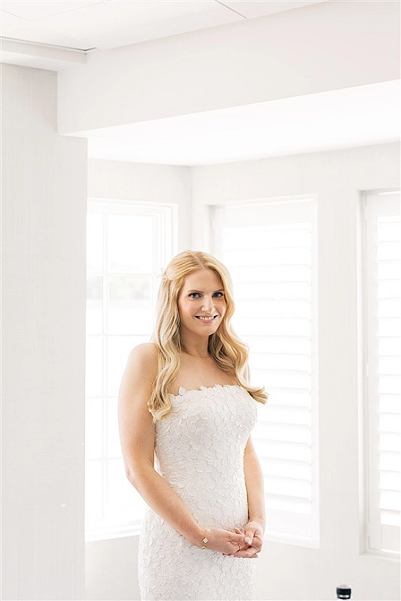 Bridal portrait of a bride in a strapless lace wedding dress with a bracelet, standing by large windows with white shutters in soft natural light